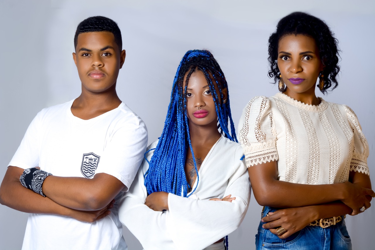 Foto de três estudantes olhando para a câmera de braços cruzados. Eles têm o semblante sério e usam camisetas claras.
