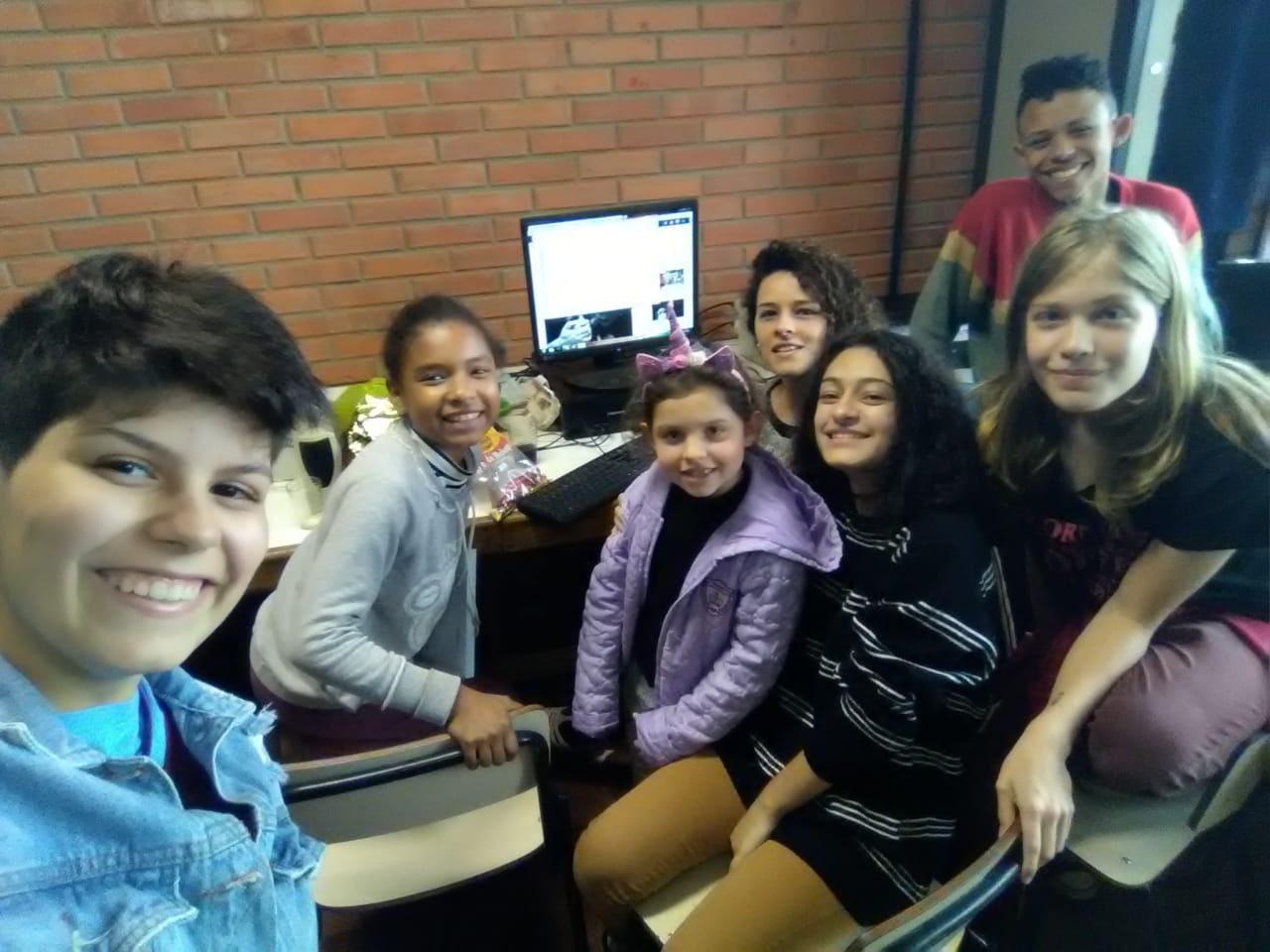 Foto com 5 estudantes e um professor ao fundo. Todos estão à frente de um computador e sorrindo para a câmera