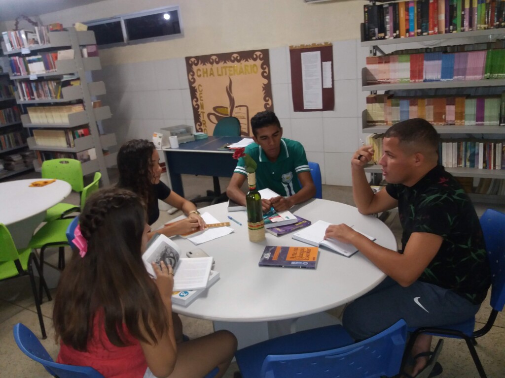 Quatro adolescentes, duas moças e dois rapazes, estão sentados ao redor de uma mesa redonda em uma biblioteca. Eles estão conversando, e todos seguram livros abertos nas mãos.