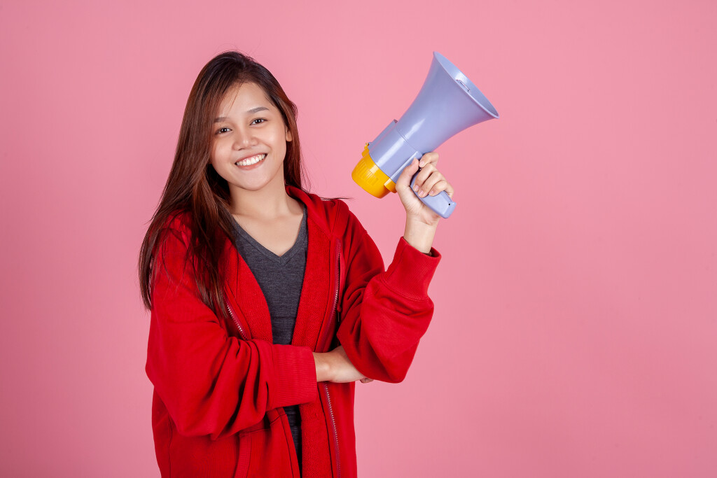Menina vestindo blusa vermelha e camiseta cinza segura um megafone e sorri